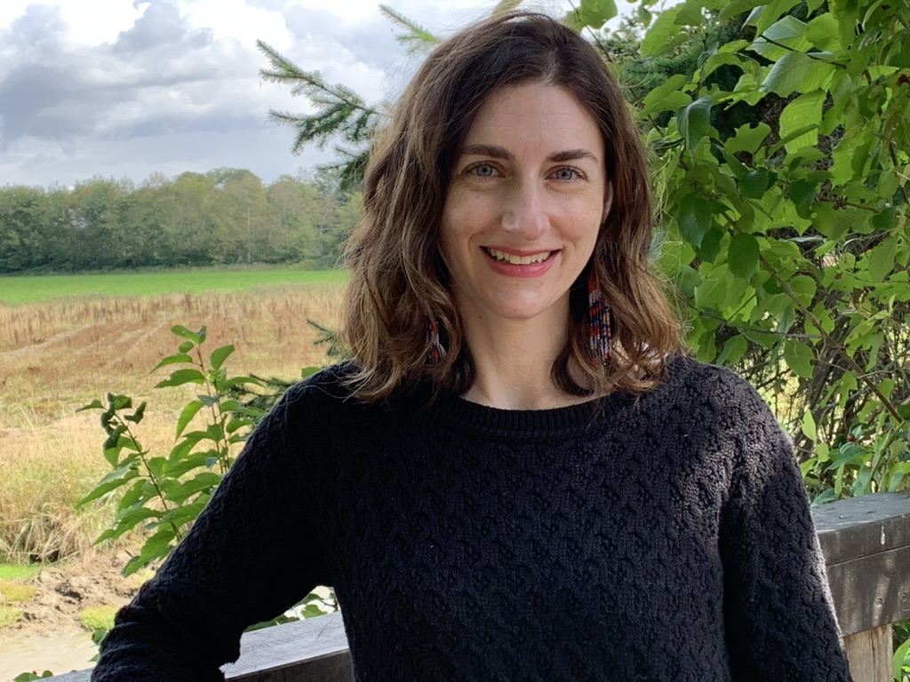 A brunette woman in a black sweater stands in front of trees and a grassy field